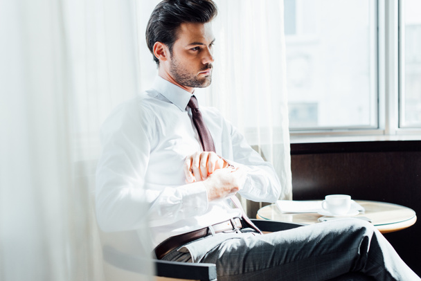 handsome bearded man in suit touching shirt while sitting near coffee table with cup of coffee - Photo, Image