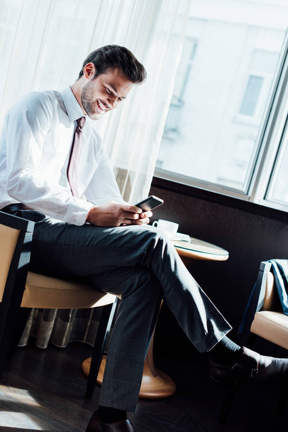 cheerful businessman in suit looking at smartphone while sitting near coffee table in hotel  - Foto, Imagem