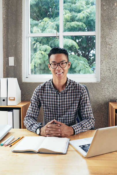 Retrato de joven atractivo hombre asiático confiado en gafas sentados en la mesa con cuaderno y sonriendo a la cámara felizmente
 - Foto, Imagen