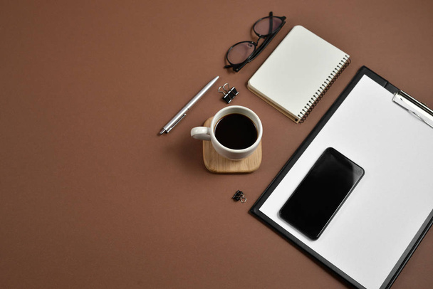 Flat lay, top view office table desk. Workspace with blank clip board, notebook, office supplies, spectacles, pen, mobile phone and coffee cup on brown background - Photo, Image