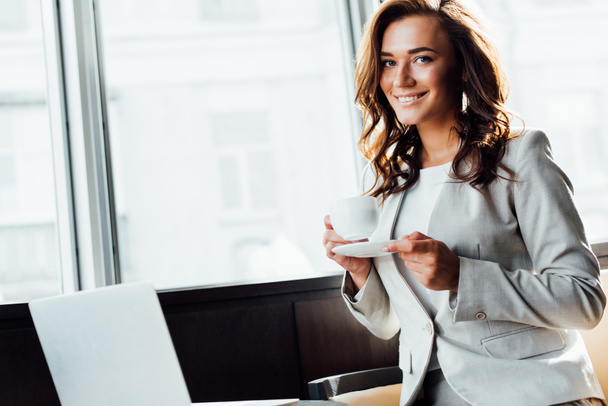 cheerful businesswoman holding cup of coffee and looking at camera - Photo, Image