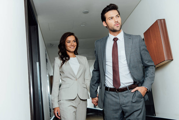 handsome businessman and happy businesswoman holding hands and walking in hotel corridor  - Foto, Imagem