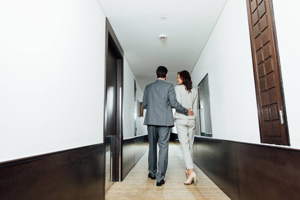 vista trasera de hombre de negocios confiado abrazando a mujer de negocios en ropa formal mientras camina en el pasillo del hotel
  - Foto, Imagen