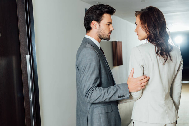 confident businessman touching hand of attractive businesswoman in formal wear while standing in hotel corridor  - Фото, зображення