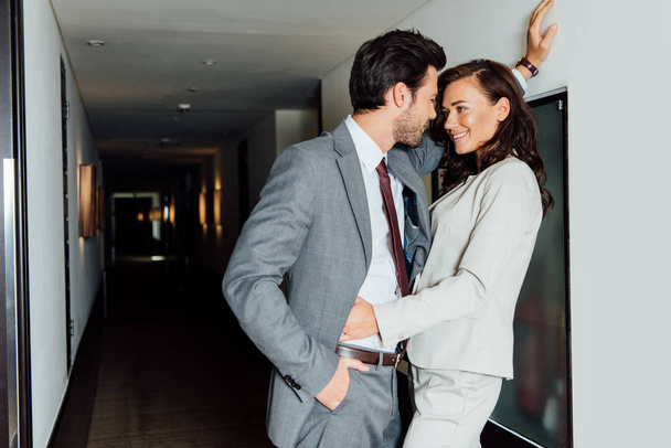 homem bonito em terno de pé com a mão no bolso e olhando para a mulher bonita no corredor do hotel
  - Foto, Imagem