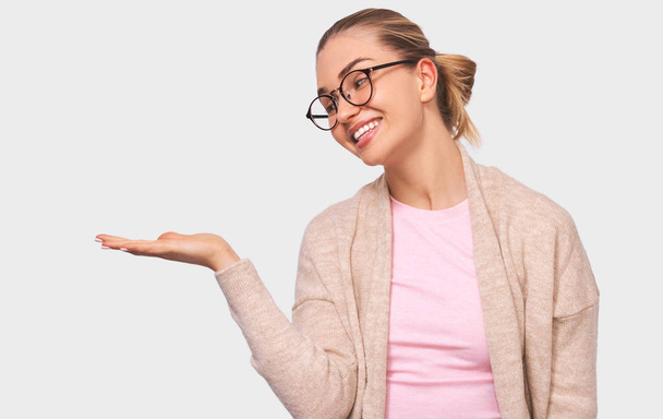 Puzzled blonde pretty young woman looks pleasant to her palm hand, showing something in her hand, wearing round transparent eyewear and casual beige and pink blouses mock up space for your advertising - Photo, Image
