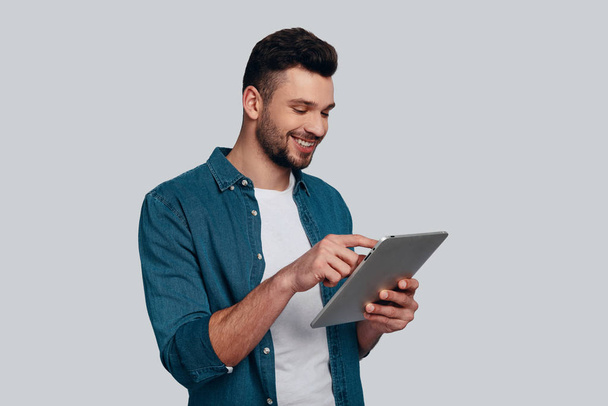 Charming young man using his digital tablet and smiling while standing against grey background - Foto, Imagem
