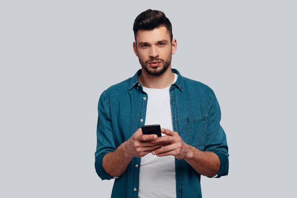 Caucasian man using his smart phone and looking at camera while standing against grey background - Fotoğraf, Görsel