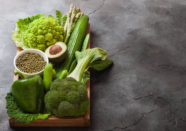 Assorted green toned raw organic vegetables in wooden box on dark background. Avocado, cabbage, cauliflower and cucumber with trimmed and mung beans and pepper and broccoli with sparagus tips. - Foto, immagini