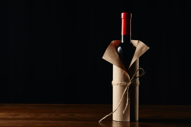 Wine bottle in paper wrapper on wooden surface isolated on black - Fotografie, Obrázek