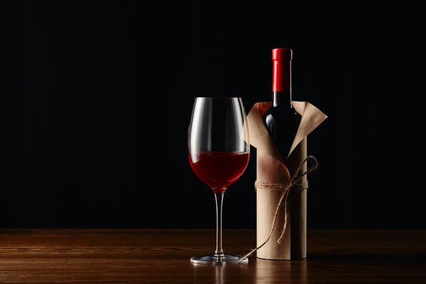 Μπουκάλι κρασί σε Xάρτινο περιτύλιγμα και το γυαλί στην ξύλινη επιφάνεια που απομονώνονται σε μαύρο - Φωτογραφία, εικόνα