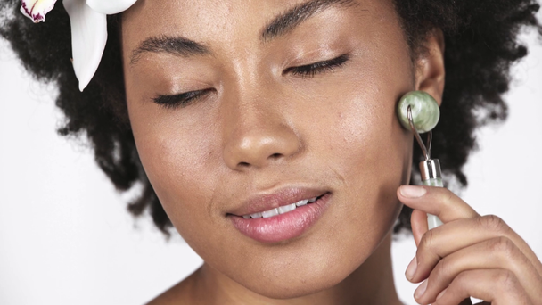 close up de atraente menina afro-americana massageando rosto limpo com rolo de jade e sorrindo isolado no branco
  - Filmagem, Vídeo