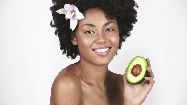 naked attractive african american girl holding and looking at half of avocado while smiling on white  - Кадры, видео