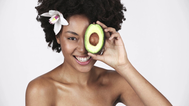 beautiful naked african american girl holding half of avocado, laughing and covering eye on white  - Footage, Video