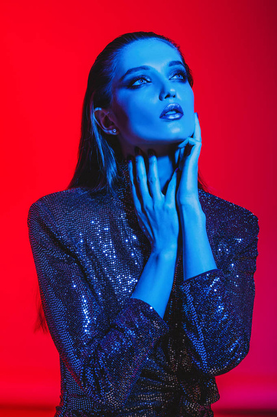 Fashion girl with long hair and stylish makeup in a black shining dress poses on the red background in neon light in the studio - Photo, image