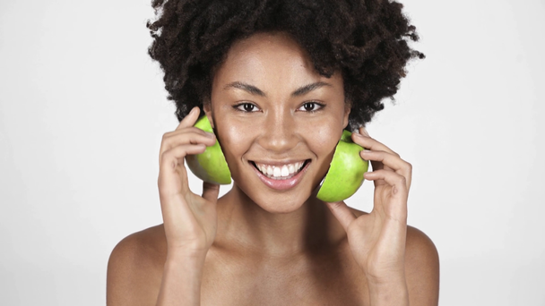 cheerful naked african american girl looking at halves of apple, applying on face and laughing on white  - Footage, Video