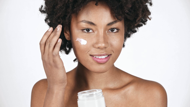 beautiful african american girl looking at container, taking face cream, applying on face and smiling on white  - Footage, Video