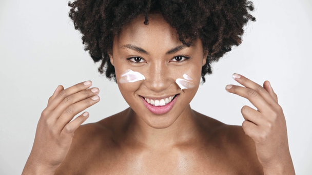 hermosa chica afroamericana mirando a la cámara aplicando crema facial en las mejillas y riendo en blanco
  - Imágenes, Vídeo