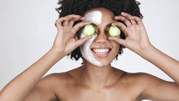 happy african american woman in in face mask covering eyes with cucumber slices on white - Metraje, vídeo