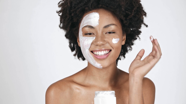 hermosa chica afroamericana en mascarilla mirando contenedor, tomando crema facial, aplicando en la mejilla y sonriendo en blanco
  - Imágenes, Vídeo