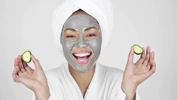 cheerful african american woman in clay mask smiling, holding slices of cucumber, covering eyes and laughing isolated on grey  - Πλάνα, βίντεο