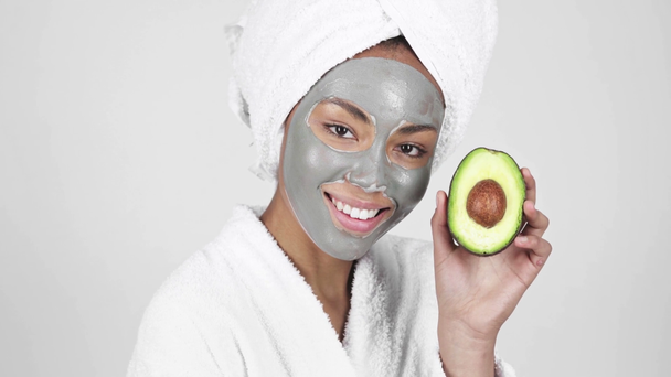 beautiful african american woman in clay mask looking at avocado and smiling isolated on grey  - Footage, Video