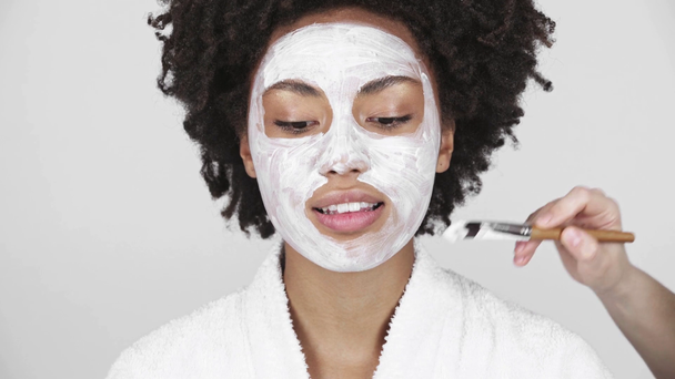 白で隔離笑顔の美しいアフリカ系アメリカ人少女の顔にマスクを適用する美容師のトリミング ビュー  - 映像、動画