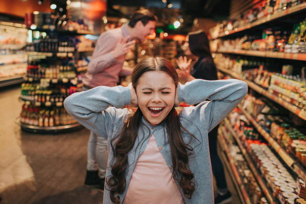 Young parents and daughter in grocery store. Girl screaming and crying. She keep ears closed. Her parents have argue and quarrel behind. - Valokuva, kuva