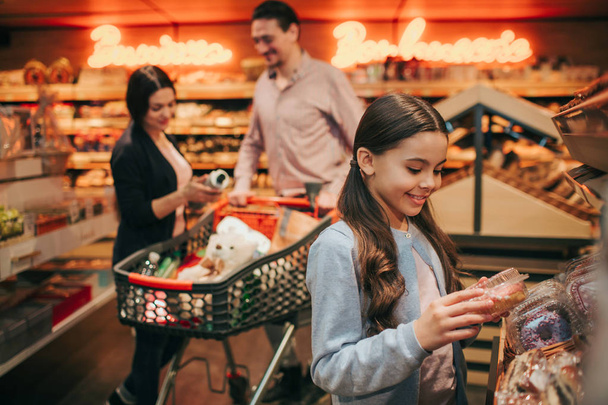 Young parents and daughter in grocery store. Cheerful small girl hold box with donut and smile. Parents stand behind at trolley with grocery. - Foto, imagen
