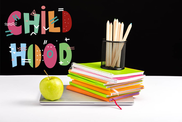 closed laptop, green apple, notebooks and color pencils on desk with childhood lettering on black  - Фото, изображение