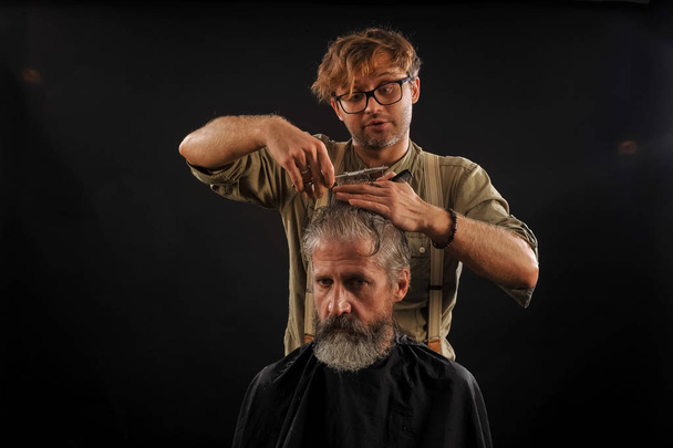 Kapper knipt senior citizen met een baard op een donkere achtergrond - Foto, afbeelding
