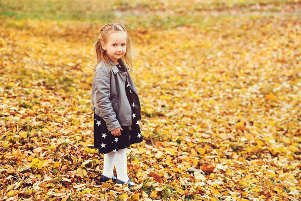 Ευτυχισμένο μικρό κορίτσι πορτρέτο σε εξωτερικούς χώρους. Φθινοπωρινό χρόνο. Μικρό ξανθό κορίτσι σε κομψό δερμάτινο παλτό και φόρεμα στο φθινόπωρο πάρκο. - Φωτογραφία, εικόνα