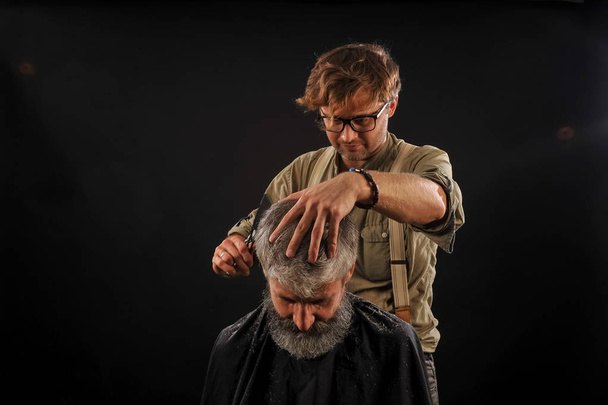 Friseur schneidet Seniorin mit Bart auf dunklem Hintergrund - Foto, Bild