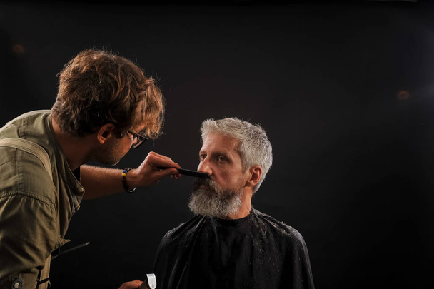 Parturi leikkaa asiakkaan parran vanhalle harmaatukkaiselle miehelle
 - Valokuva, kuva