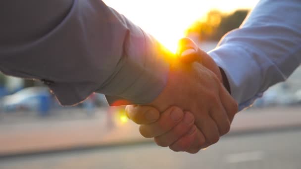 Two successful businessmen greeting each other against the background of cars parking. Young colleagues meeting and shaking hands at city street on sunset. Business handshake outdoor. Slow motion - Footage, Video