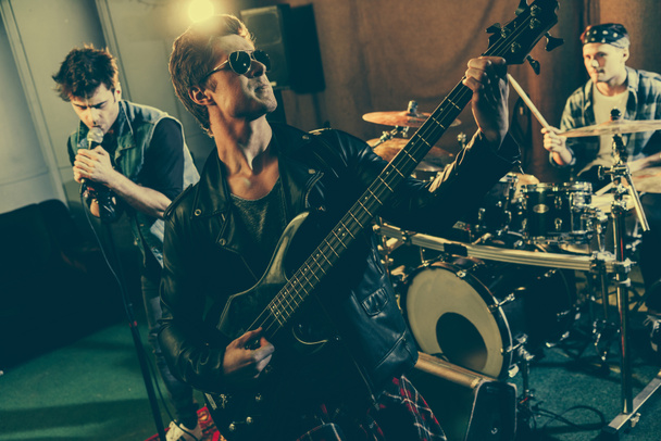 επιλεκτική εστίαση του όμορφος κιθαρίστα σε γυαλιά ηλίου παίζοντας ηλεκτρική κιθάρα κοντά ροκ μπάντα  - Φωτογραφία, εικόνα