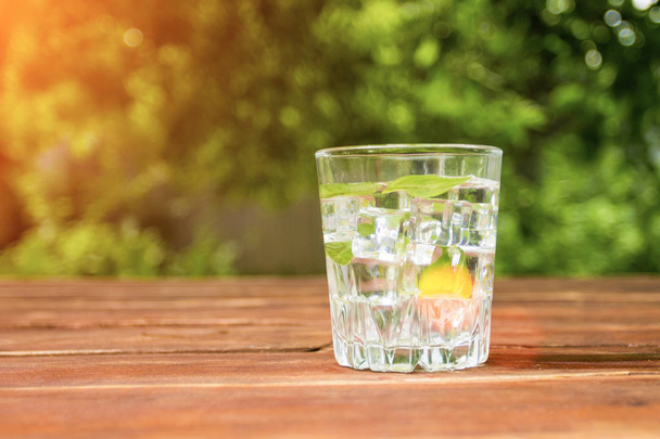 ein Glas mit einem kalten Erfrischungsgetränk mit Eis und Minzblättern auf einem Holztisch und im Hintergrund des Gartens. Konzept der Freizeitgestaltung im Freien - Foto, Bild