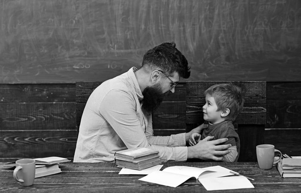 Male teacher explaining something to the boy. Side view bearded man in glasses and blond child in front of green board. Teacher holding naughty kid - 写真・画像