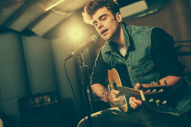 handsome singer singing in microphone while playing acoustic guitar  - Foto, afbeelding