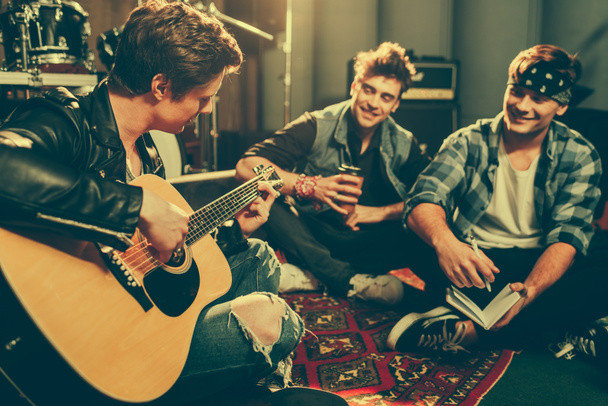 selective focus of musician playing acoustic guitar near happy friends sitting with crossed legs - 写真・画像