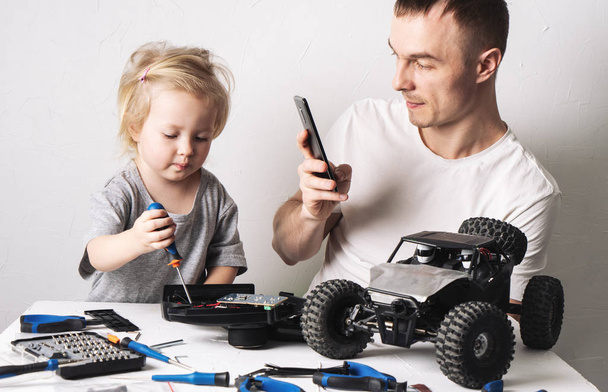 Family time: Daughter with father repairs a radio-controlled buggy model. Dad shoots a repair process on his smartphone. - Foto, Imagen