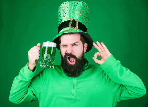 The best beer is irish. Bearded man toasting to saint patricks day. Hipster in leprechaun hat holding beer mug. Celebrating saint patricks day in bar. Irish man gesturing ok sign to green beer - Фото, изображение