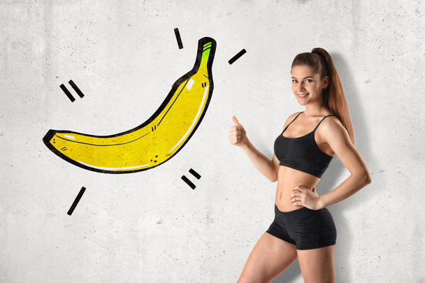Young fit woman in black gym crop top and shorts standing in half-turn doing thumbs up pointing at drawing of big banana on wall. - Foto, Bild