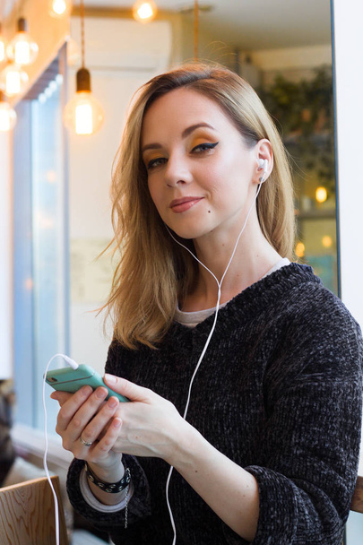 beautiful smiling young woman in gray sweater using mobile phone with earphones in cafe - Фото, изображение