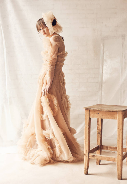 Full length portrait of beautiful young woman in nude tulle dress and tulle hair accessory, standing near wooden chair. Fabric on background. Dreamy mood - Φωτογραφία, εικόνα
