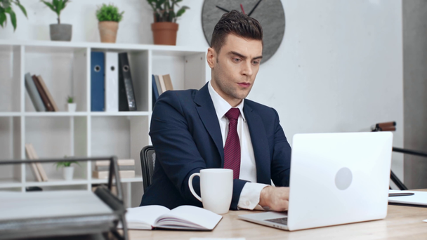 hombre de negocios guapo usando el ordenador portátil, mirando el cuaderno, beber café y escribir en portapapeles mientras está sentado en el lugar de trabajo en la oficina
 - Imágenes, Vídeo