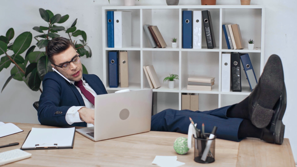 knap, ernstige zakenman op werkplek zitten met gekruiste benen op Bureau, met behulp van laptop, praten over smartphone en koffie drinken - Video