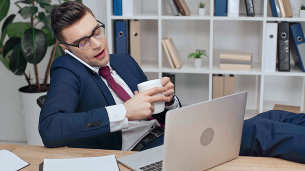 όμορφος, σοβαρός επιχειρηματίας χρησιμοποιώντας φορητό υπολογιστή, μιλώντας στο smartphone και πίνοντας καφέ ενώ κάθεται στο χώρο εργασίας με τα πόδια στο γραφείο - Πλάνα, βίντεο