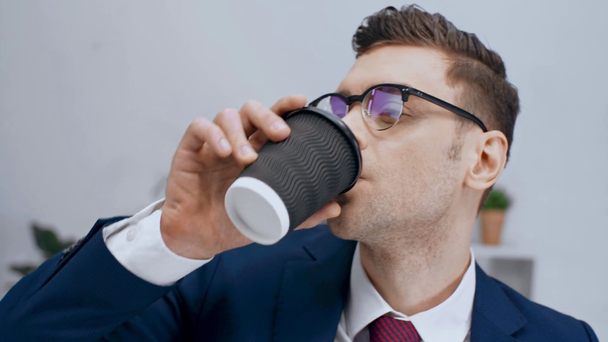 handsome businessman in eyeglasses looking at camera, smiling and drinking coffee from disposable cup - Footage, Video