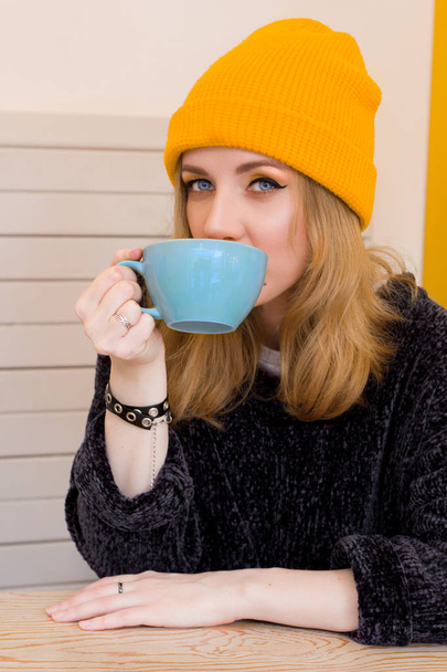 beautiful dreamy woman in a yellow hat sitting at a table in a coffee shop with blue cup of warming drink - Valokuva, kuva
